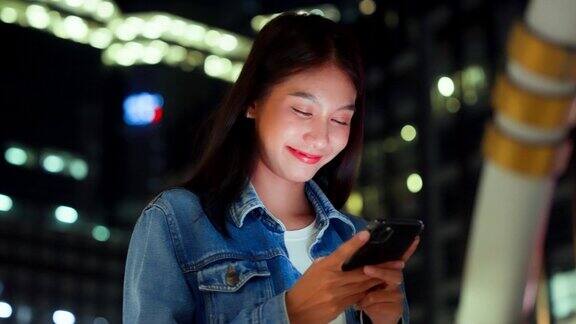 美丽的年轻亚洲女子在晚上用手机走在城市街道上一个快乐的女人站在那里用手机打字