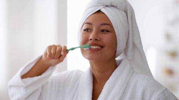 早晨例行公事年轻开朗的亚洲妇女穿着浴衣和毛巾在浴室刷牙