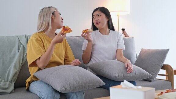 两个年轻的亚洲女性朋友欢快地吃着披萨看着电视坐在家里客厅的沙发上亚洲休闲用餐