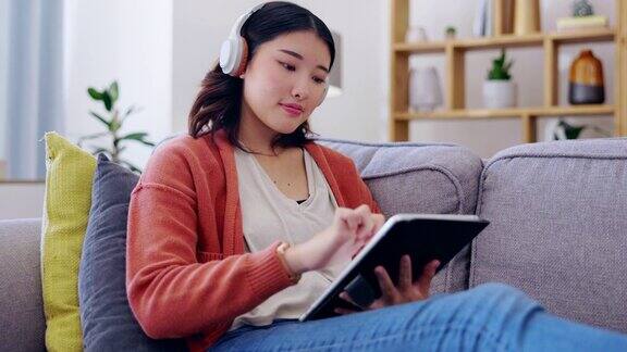 平板电脑耳机和亚洲女人在沙发上的音乐流媒体应用程序在线游戏和创造性的e学习在家里z世代或年轻的韩国女孩在沙发上放松数码科技和听音频