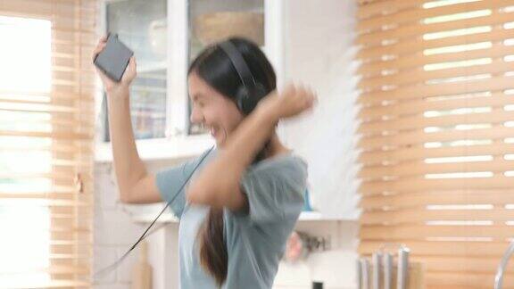 年轻美丽的亚洲女人在厨房跳舞同时拿着智能手机在家里听音乐早上