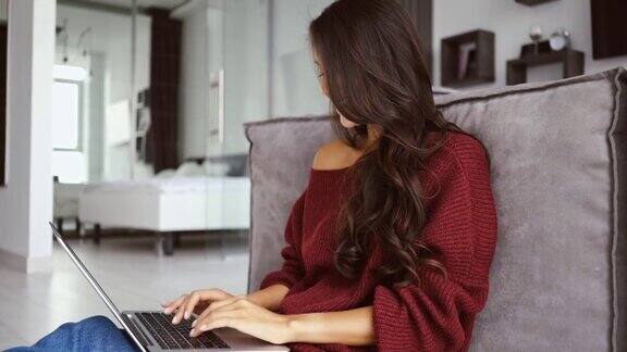 面带微笑的黑发女人在毛衣坐在地板上使用笔记本电脑
