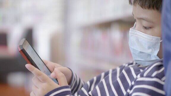 亚洲小男孩在图书馆学习在线手机戴口罩预防冠状病毒covid-19概念技术教育