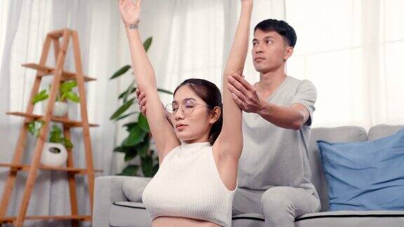 4k美丽的亚洲孕妇坐在地板上做瑜伽姿势她的丈夫把她的手臂举到背后在房子的客厅安全男人在后面照顾妻子