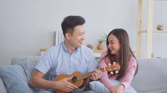 年轻浪漫的亚洲夫妇在客厅的沙发上玩尤克里里亚洲男朋友唱歌和演奏尤克里里为美丽的女朋友放松在沙发上在家里