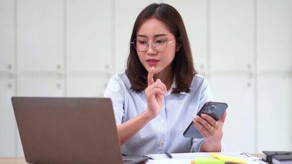 亚洲女商人一边在办公室用笔记本电脑工作一边使用智能手机坐在办公桌前查看手机
