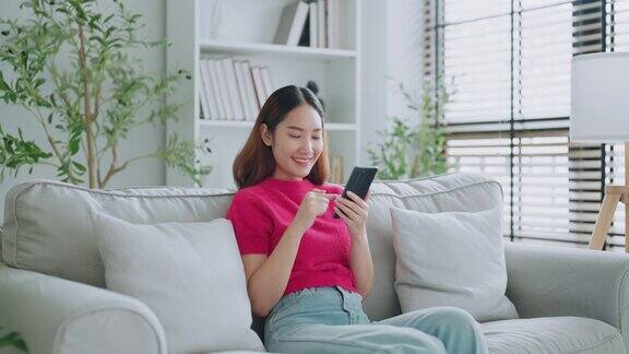 快乐的年轻亚洲女人在家里舒适的沙发上放松地用智能手机发短信微笑的女孩在家里用手机聊天网上购物视频通话交流