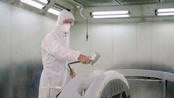 图为一名穿着白色PPE防护服的白人男子在汽车保险杠上喷涂底漆送修车时用心、细致地修理油漆