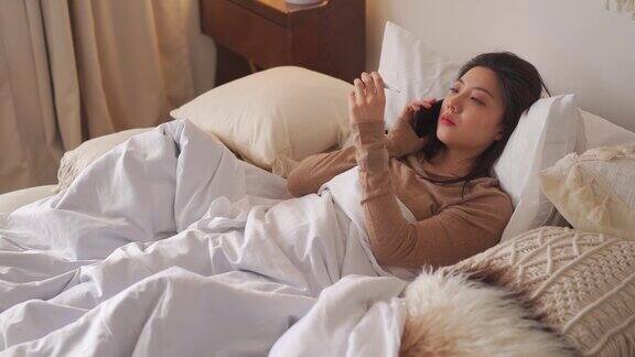 一名亚洲华裔妇女躺在床上盖着毯子患感冒和流感用智能手机和朋友聊天用数字体温计测量体温