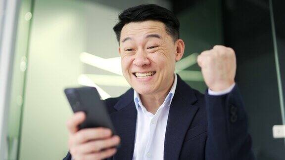 关闭了亚洲商人坐在现代化的办公室里用智能手机阅读好消息微笑着庆祝成功