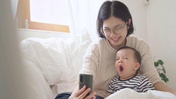 微笑的亚洲母亲和她可爱的儿子在家里使用智能手机小儿子打哈欠
