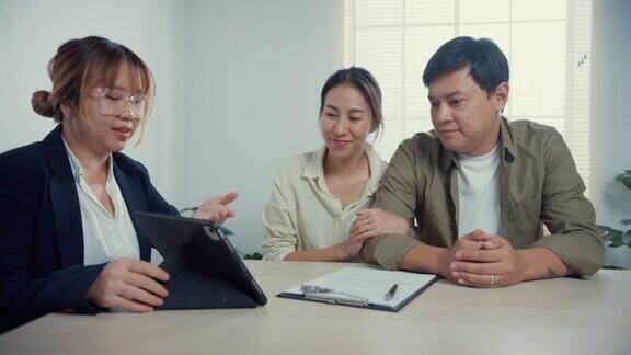 女销售员不停地向年轻的亚洲夫妇解释房产细节他们随意地坐在书桌前对房产办公室的新房合同协议感到满意
