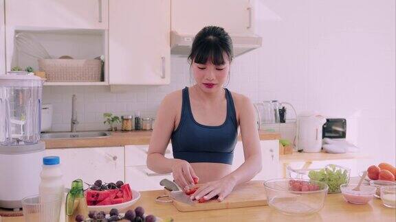 一个穿着运动服的亚洲女人开心地切蔬菜把西红柿放在玻璃碗里做沙拉她在厨房里拥抱快乐和健康的生活方式通过营养丰富的烹饪促进健康