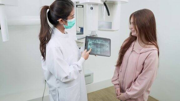 在x光室一名带着防护面罩的亚洲女牙医正在向一名白人少女病人解释用药片拍摄的放射照片而她正在仔细地听着