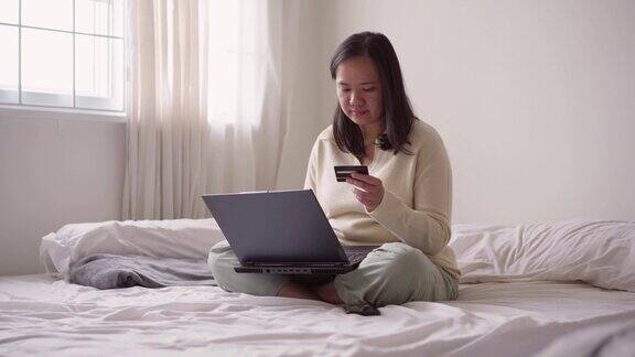快乐的年轻亚洲妇女坐在床上用信用卡和笔记本电脑在线支付