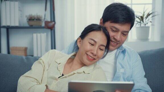 年轻的亚洲夫妇休闲地坐在沙发上使用平板电脑在线购物在家里的客厅观看在线娱乐电影