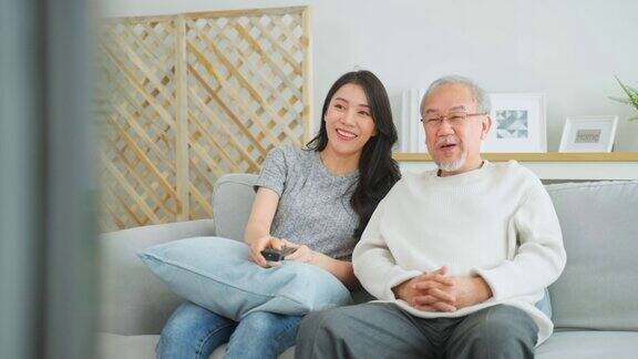 亚洲年长的父亲和女儿一起在家里看滑稽电影慈祥的老爷爷和美丽的女孩孙女坐在沙发上欣赏电视上的喜剧节目在客厅里度过时光