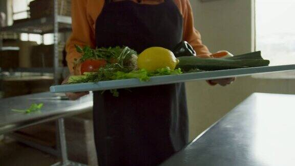 一位年长的白人妇女拿起一个装满蔬菜的切菜板把它放在一个商业厨房的不锈钢台面上