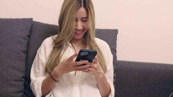近景:穿着白衬衫的亚洲女性坐在沙发上用智能手机在客厅浏览社交媒体