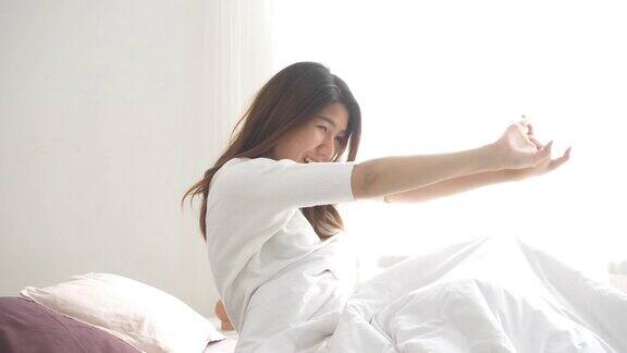 幸福美丽的年轻亚洲女人早上醒来坐在床上伸展在舒适的卧室看着窗外有趣的亚洲女人醒来后亚洲妇女正在伸展和微笑