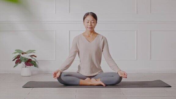 平静的健康亚洲年轻女性坐在地毯上呼吸瑜伽莲花姿势瑜伽冥想年轻健康女性放松和舒适的白色舒适的家瑜伽锻炼为健康