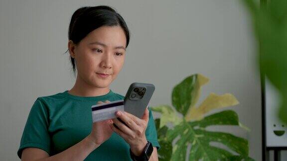 亚洲女性手持信用卡在家办公室用智能手机进行网上支付