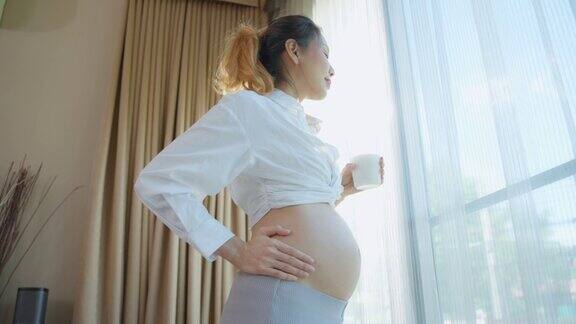 亚洲美女孕妇喝一杯奶养生迷人的孕妇站在客厅的室内用幸福和爱抚摸着子宫里的婴儿