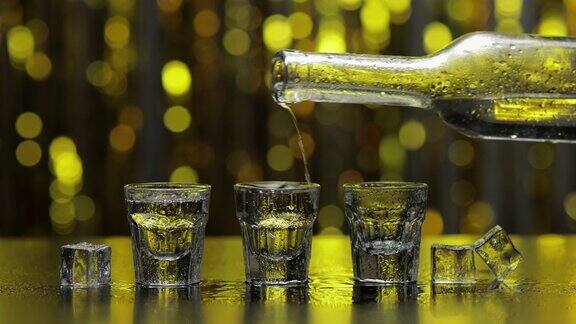 酒保将冰冻的伏特加从瓶子倒入小酒杯冰块映衬着闪亮的金色派对背景