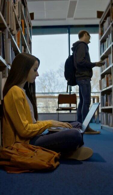 SLOMO女子使用笔记本电脑而其他学生在图书馆的书架上找一本书