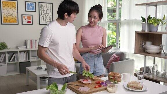 年轻快乐的亚洲夫妇在家里的厨房里用平板电脑微笑着做饭