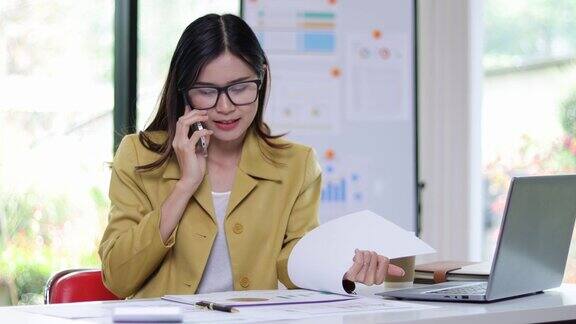 一位女商人正在给客户打电话她在读办公室桌上的一些文件一个女人一边和别人打电话一边分析文件和阅读每月的工作计划