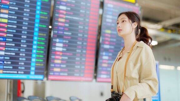 年轻的亚洲妇女拿着护照和登机牌在国际机场看航班信息板检查她的航班