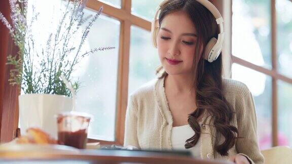 亚洲自由职业女性成人的微笑肖像一边用平板电脑远程在线工作一边听着耳机里的音乐心情愉悦积极向上在咖啡馆餐厅里自然光透过橱窗商店咖啡馆