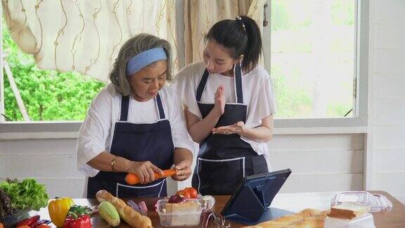 快乐的亚洲小女儿和年长的母亲在平板电脑上烹饪在线课程一起在家里的厨房里做新鲜的蔬菜食物