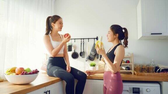 亚洲活跃的两个女人姐妹穿着运动服在厨房吃苹果年轻漂亮的女孩妹妹心情愉快喜欢吃水果健康的食物来减肥和保健在家里
