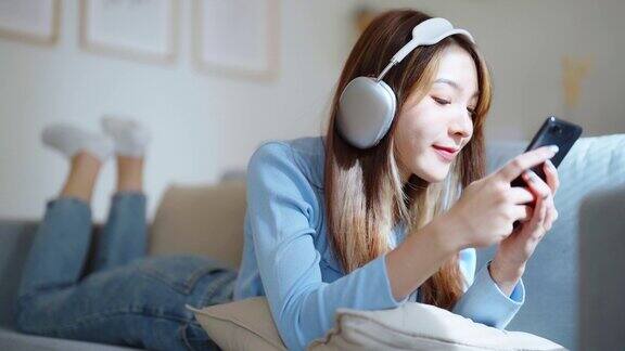 快乐的年轻亚洲女子在家里放松女性微笑着坐在沙发上手里拿着智能手机女孩用视频和朋友通话听音乐