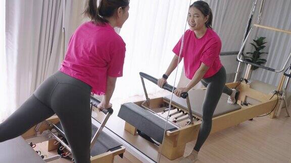 运动亚洲妇女在突出的颜色衬衫伸展她的腿通过普拉提改革机器在镜子前