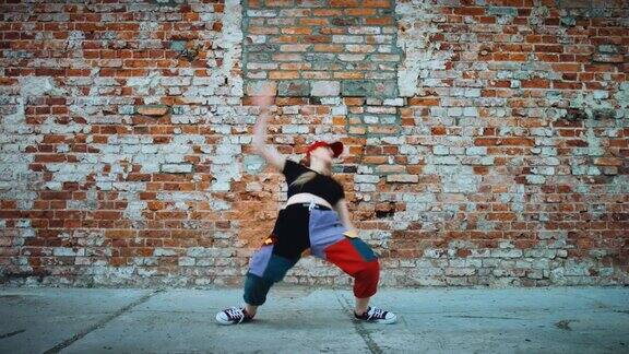一名兴高采烈的年轻女子戴着一顶帽子活跃地在城市砖墙旁边的街道上跳舞穿着五颜六色的裤子127bpm股票视频