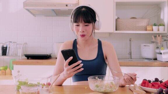 一个穿着运动服的亚洲女人一边玩手机一边愉快地品尝着蔬菜沙拉她喜欢吃有益的食物在她的厨房里提倡快乐和健康的生活方式