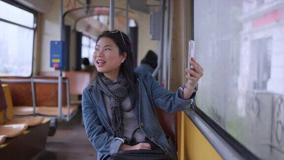 快乐的年轻亚洲女性穿着休闲服装用智能手机与朋友视频通话微笑着坐在欧洲观光巴士上女子在维也纳旅游