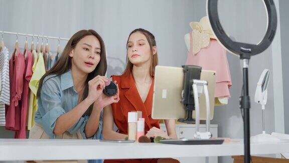 亚洲两名女性视频博主对着镜头回顾在家里的美容美妆博主网红女性朋友在办公室商店的平板电脑上直播并向粉丝展示化妆粉