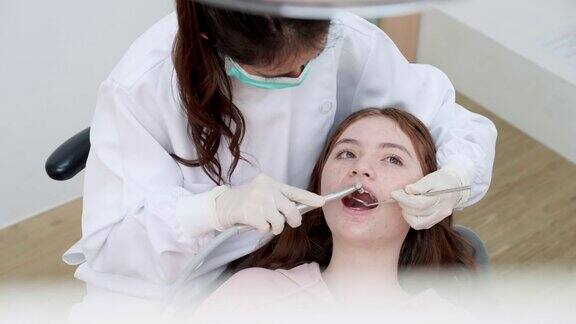 亚洲中年女牙医为十几岁的少女患者做牙科检查和补牙使用角度镜、吸唾液等牙科设备少女坐在牙科椅上高角度观看