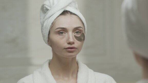 在家庭盥洗室里一个裹着毛巾的女人对着镜子敷眼睛下面的眼罩