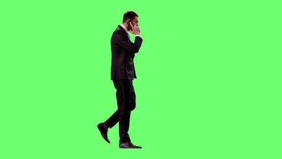 一个穿着西装的商人正在绿色屏幕的背景下通过移动智能手机边走边说话