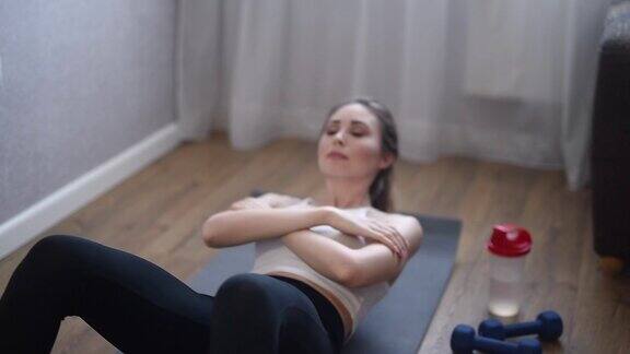 一个年轻女子在家里的地板上做运动她在瑜伽垫上摇晃她的腹部运动健康的生活方式在前景的哑铃