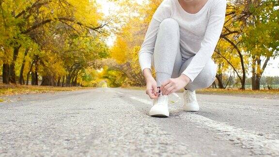 跑步者系着鞋带在柏油路上奔跑在秋天的大自然中年轻的女子准备开始跑步女孩从事户外运动概念健康的生活方式和身体护理