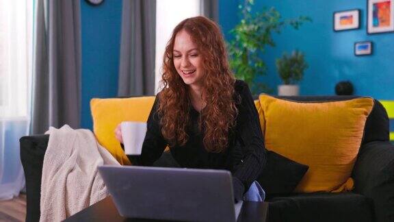 微笑的白人少女学习笔记本电脑而坐在沙发沙发在时尚舒适的客厅快乐女孩在家里玩得开心上网用杯子喝咖啡