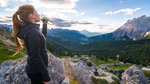 日落时分一个女人在山顶上喝着水瓶里的水俯瞰着山谷