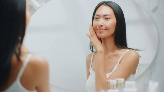 美丽的亚洲女人扮演和欣赏她完美柔软的皮肤和繁茂的黑发看着相机和微笑在浴室的镜子健康自然美容有机护肤品