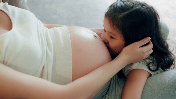 怀孕的母亲和学龄前的女儿坐在家里的沙发上可爱的女孩亲吻怀孕妈妈的肚子